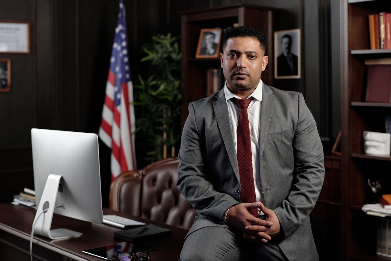 A professional businessman in a suit sitting in an office setting, with the American flag in the background.